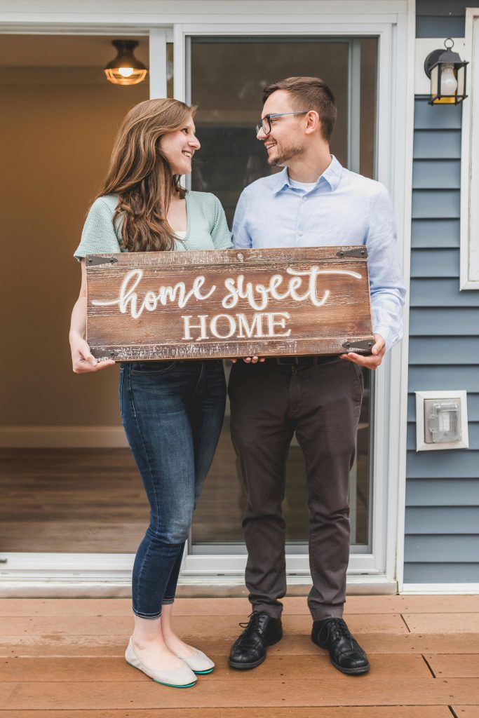 Young couple standing outside new home, holding 'home sweet home' sign, joyful and smiling.
